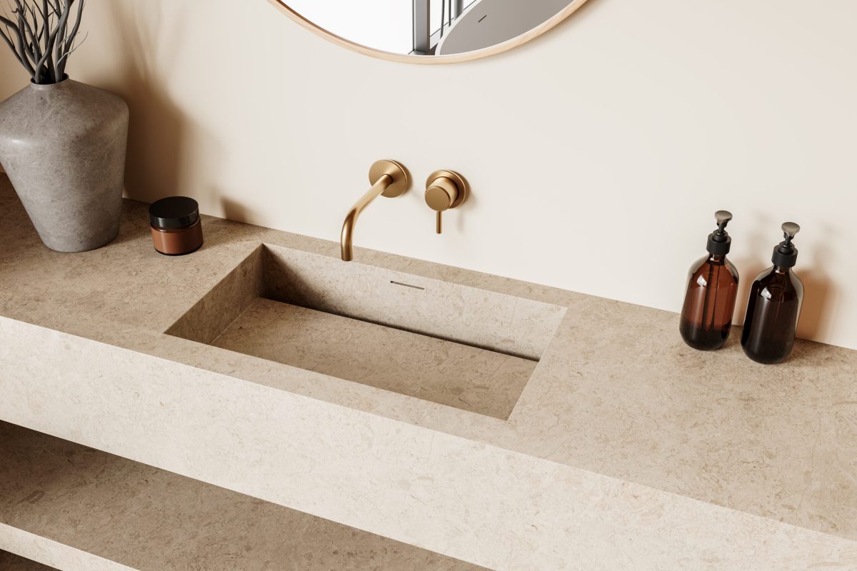 Badezimmer mit beigem Lavabo aus Stein mit goldenen Wandarmaturen und rundem Spiegel.